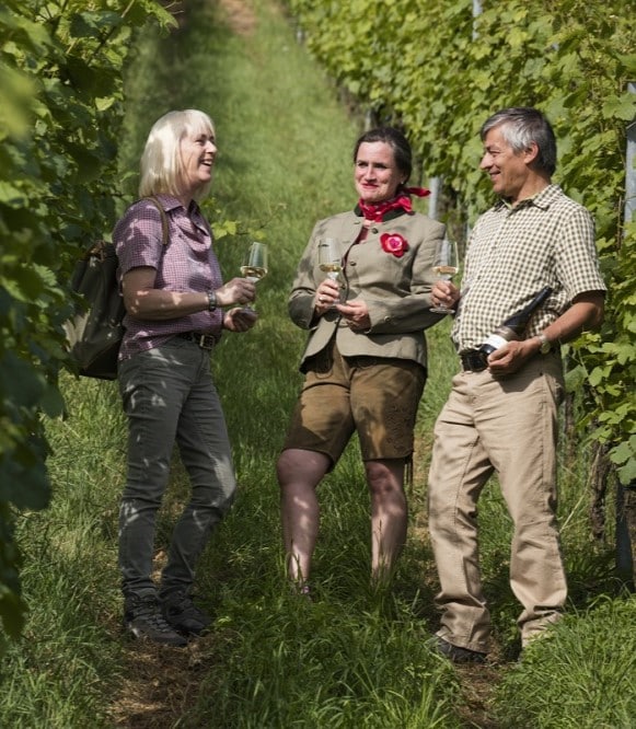 Ein Mann und zwei Frauen stehen in den Weinbergen und trinken ein Glas Wein.